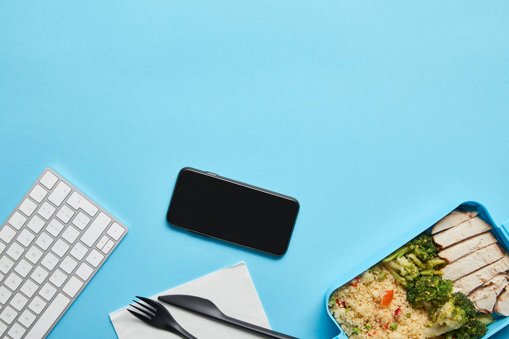 Vista dall'alto del posto di lavoro con smartphone, tastiera del computer e lunch box con risotto, pollo e broccoli su sfondo blu
 - Foto, immagini
