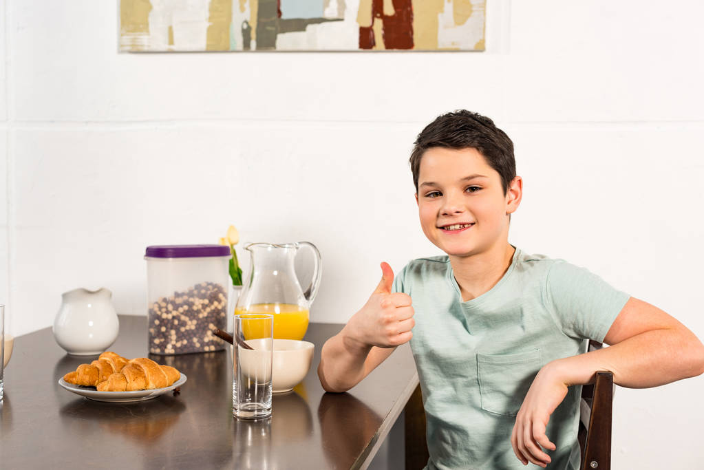 niño sonriente sentado en la mesa durante el desayuno y mostrando el pulgar hacia arriba
 - Foto, Imagen