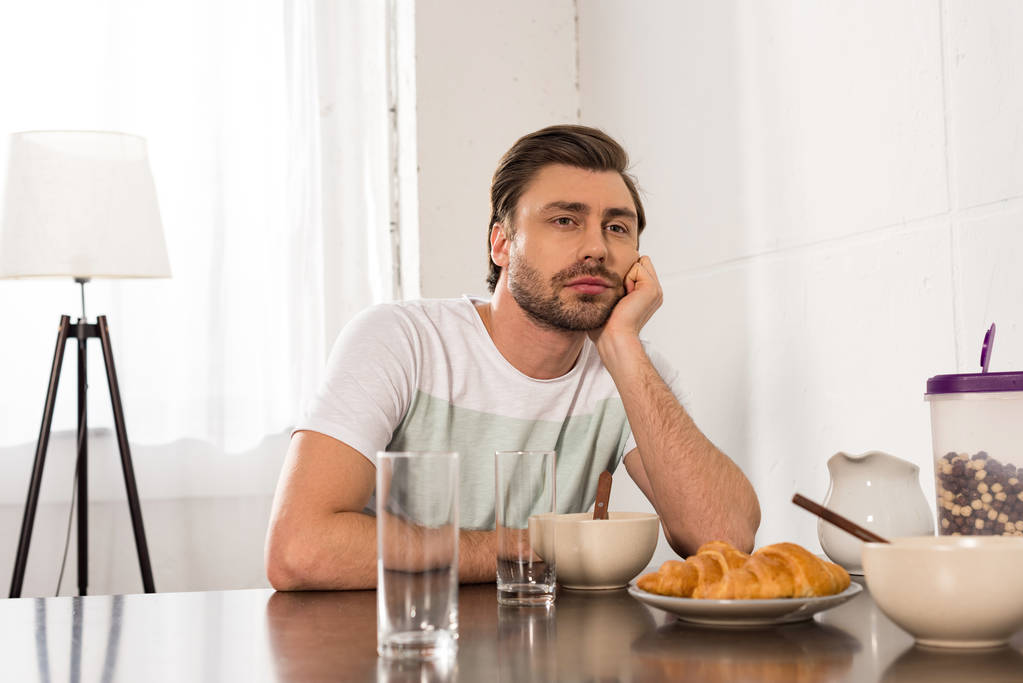 σκεπτικός άνθρωπος κάθεται στο τραπέζι και με τα μούτρα με το χέρι κατά τη διάρκεια του πρωινού στην κουζίνα - Φωτογραφία, εικόνα