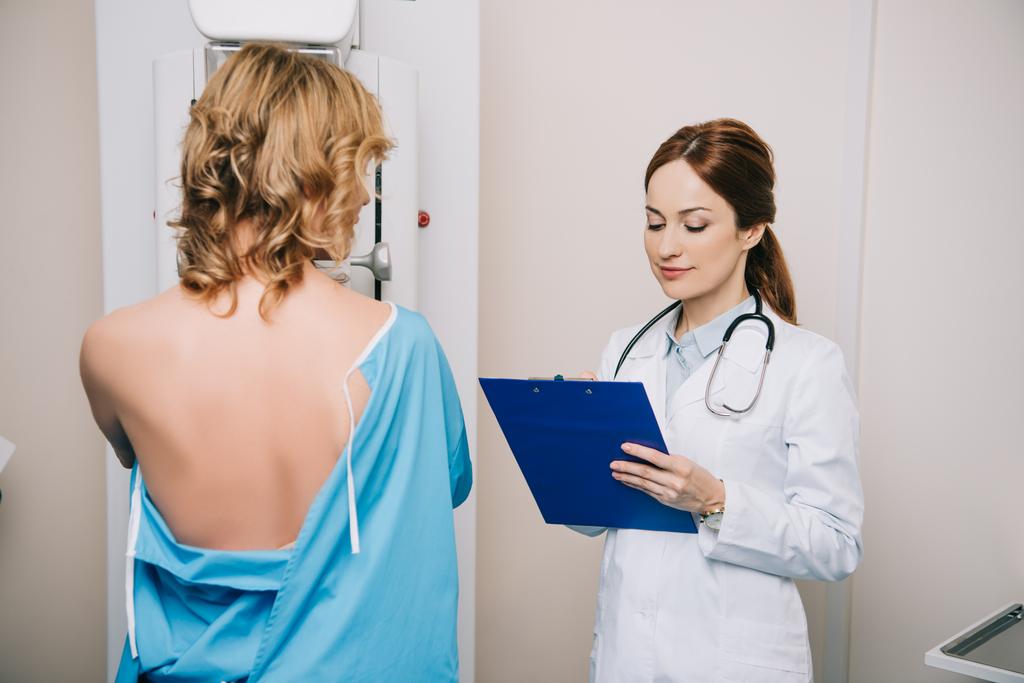 hermoso médico joven mirando el portapapeles mientras está de pie cerca del paciente durante la prueba de mamografía en la máquina de rayos X
 - Foto, imagen