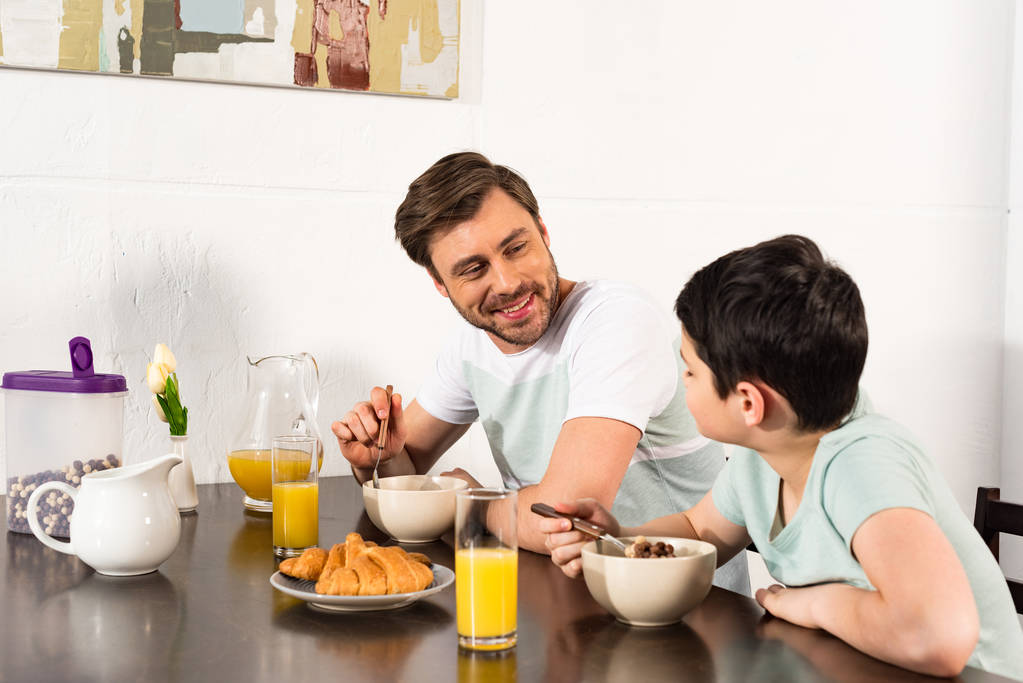 hymyilevä isä ja poika aamiaisella ja katsomalla toisiaan keittiössä
 - Valokuva, kuva