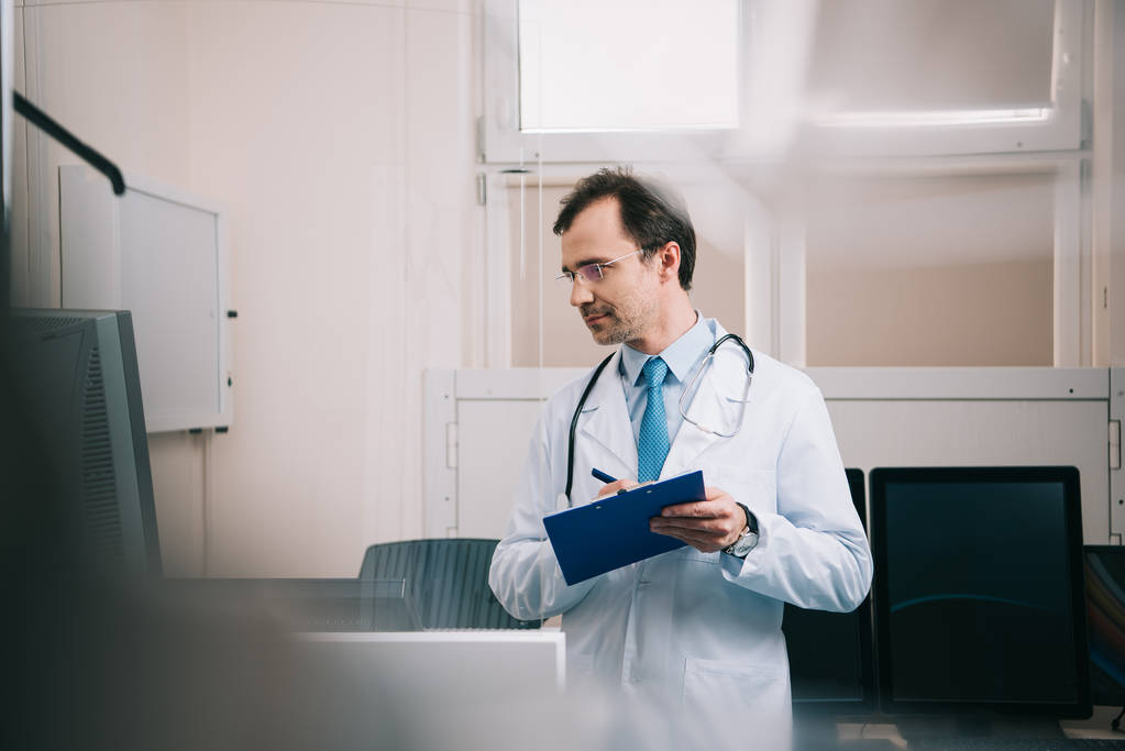 selective focus of smiling doctor writing on clipboard and looking away  - Photo, Image