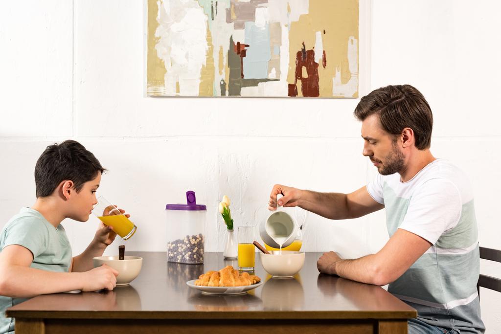Vater schüttet Milch in Schüssel, während Sünde beim Frühstück Orangensaft trinkt - Foto, Bild