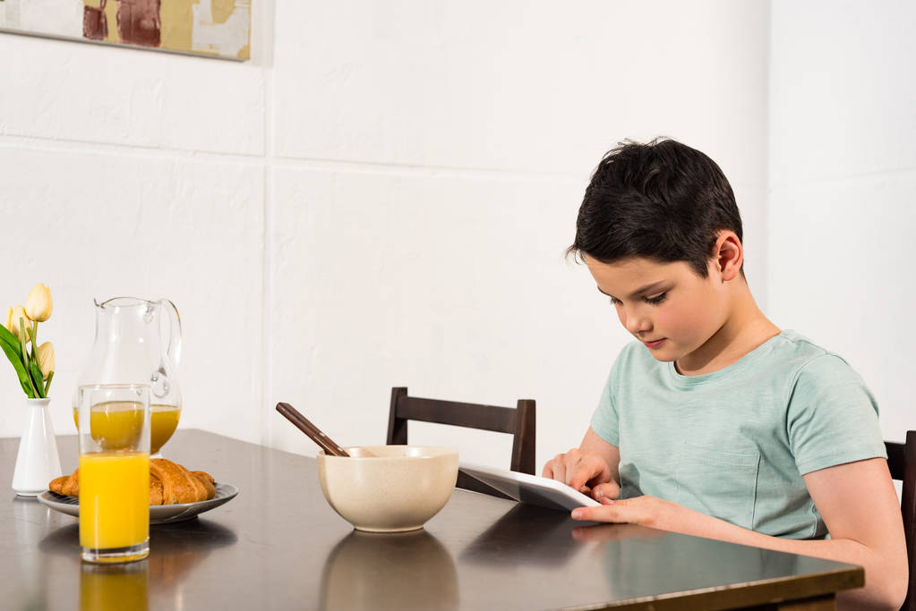 niño usando tableta digital durante el desayuno en la cocina
 - Foto, Imagen