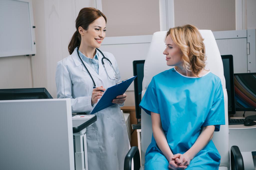 veselý lékař drží podložku v blízkosti krásné ženy sedí v křesle v nemocnici - Fotografie, Obrázek