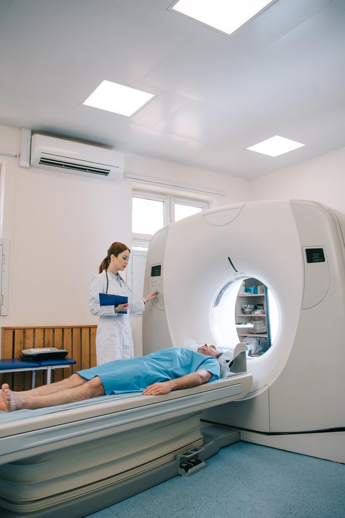 Radiologe im weißen Kittel operiert CT-Scanner, während Patient auf CT-Bett liegt  - Foto, Bild