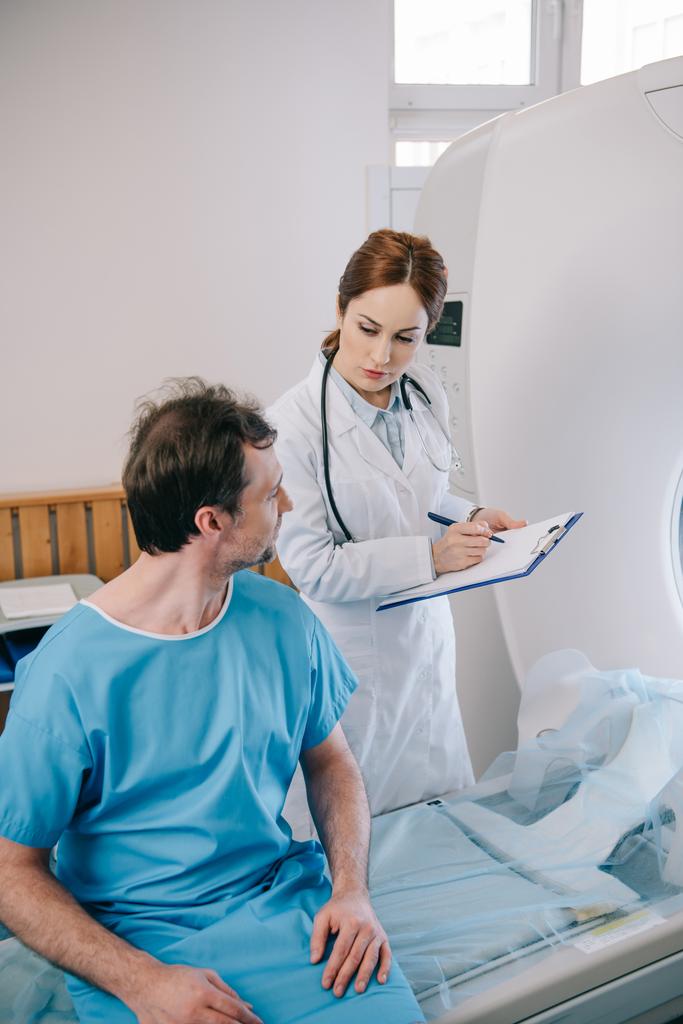 ctスキャナーベッドの上に座っている患者の近くに立っている間、クリップボードに書き込む注意深い医師 - 写真・画像
