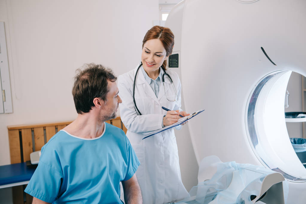 uśmiechnięty radiolog piśmie na Schowka stojąc w pobliżu pacjenta siedzi na CT skanera łóżko - Zdjęcie, obraz