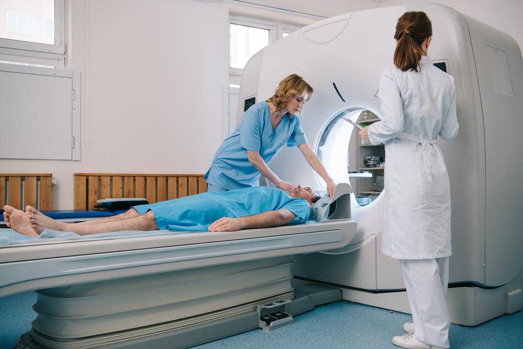 Ακτινολόγος σε λευκό παλτό στέκεται κοντά στο CT σαρωτή, ενώ η νοσοκόμα προετοιμάζει τον ασθενή για διαγνωστικούς ελέγχους - Φωτογραφία, εικόνα