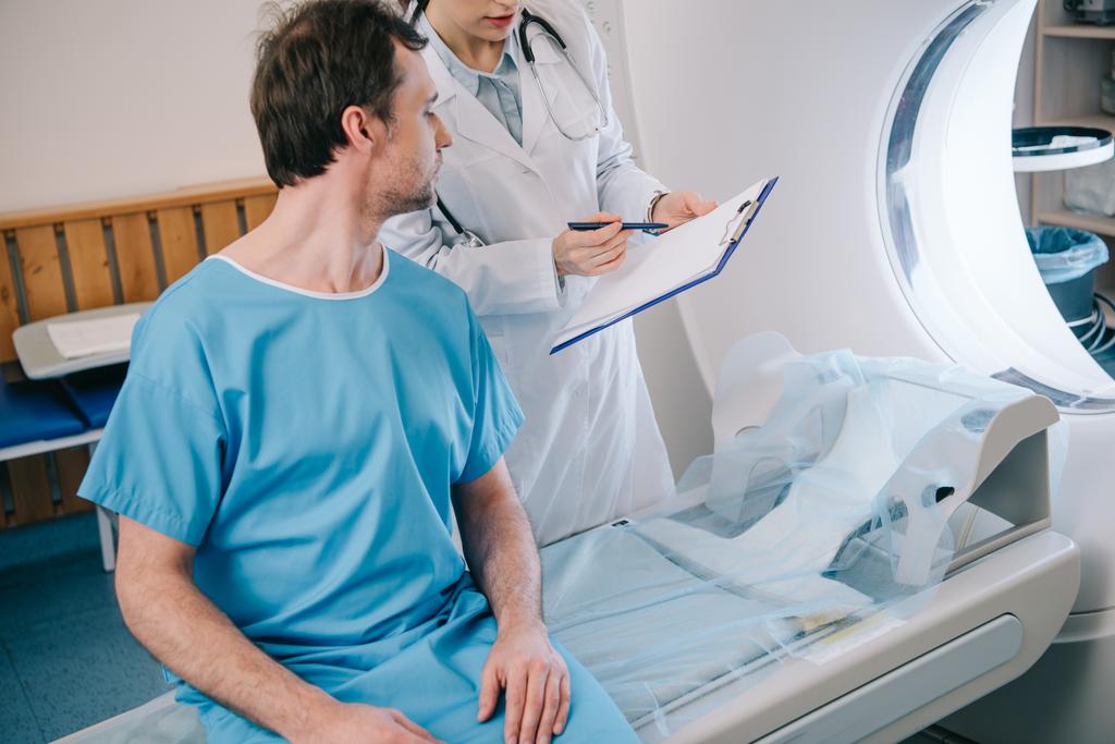 ctスキャナーベッドに座っている患者に診断を示す若い放射線科医の部分的な眺め - 写真・画像