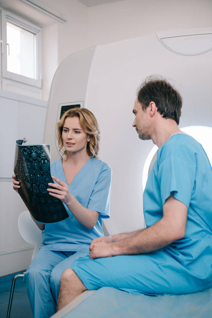 piękny lekarz patrząc na diagnostyki rentgenowskiej w pobliżu pacjenta siedzi na MRI skanera łóżko - Zdjęcie, obraz
