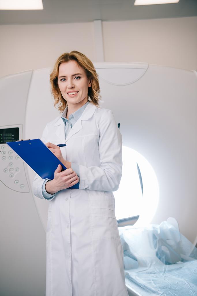 uśmiechnięty radiograf piśmie na Schowka i patrząc na kamerę stojąc w pobliżu MRI maszyny  - Zdjęcie, obraz