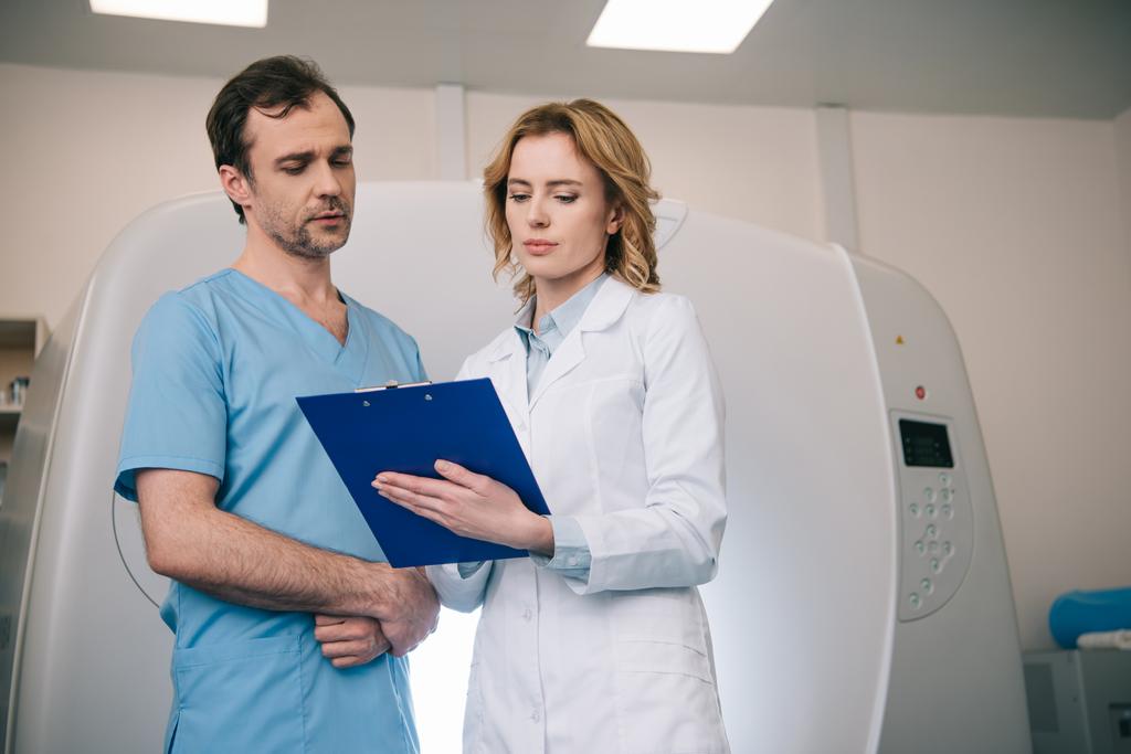 zwei Radiologen, die in der Nähe eines Computertomographen die Diagnose auf dem Klemmbrett betrachten - Foto, Bild