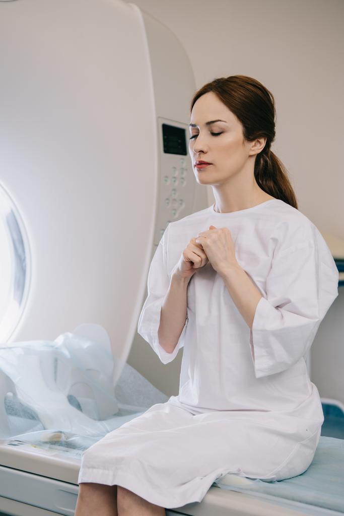 病院のmriスキャナーベッドに座りながら、目を閉じて祈る魅力的な女性 - 写真・画像