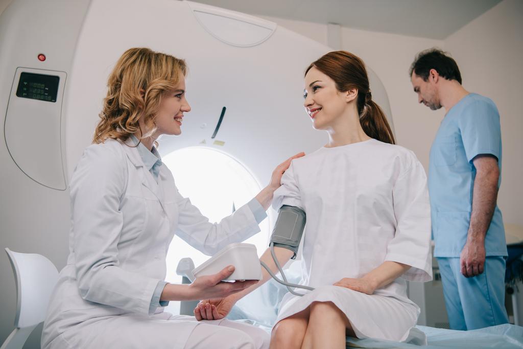 улыбающийся доктор измеряет пациентам артериальное давление тонометром, а ассистент регулирует КТ сканер
 - Фото, изображение
