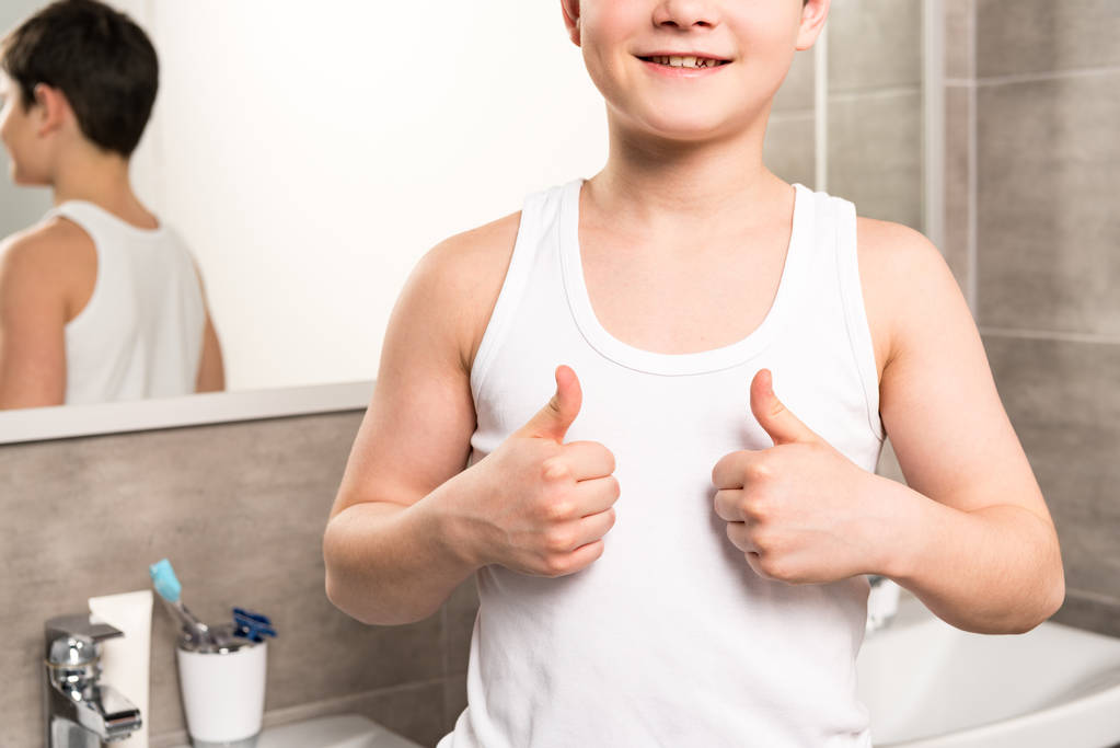 bijgesneden weergave van lachende jongen die duimen omhoog in de badkamer - Foto, afbeelding