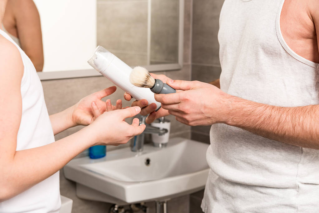 cropped view of dad giving shaving foam and shaving razor to son in bathroom - Photo, Image