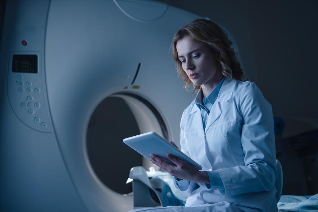 médico serio que mira la tableta digital con diagnóstico de rayos X mientras está sentado cerca del escáner ct
  - Foto, Imagen