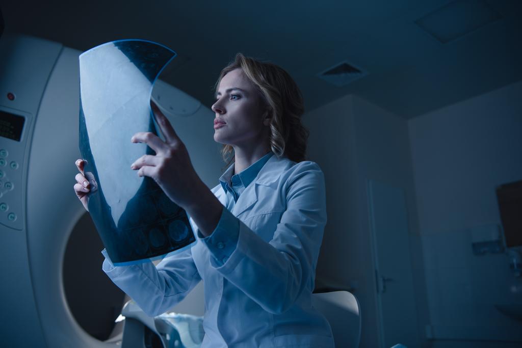 Schöne nachdenkliche Ärztin betrachtet Röntgendiagnose, während sie in der Nähe des Computertomographen sitzt - Foto, Bild