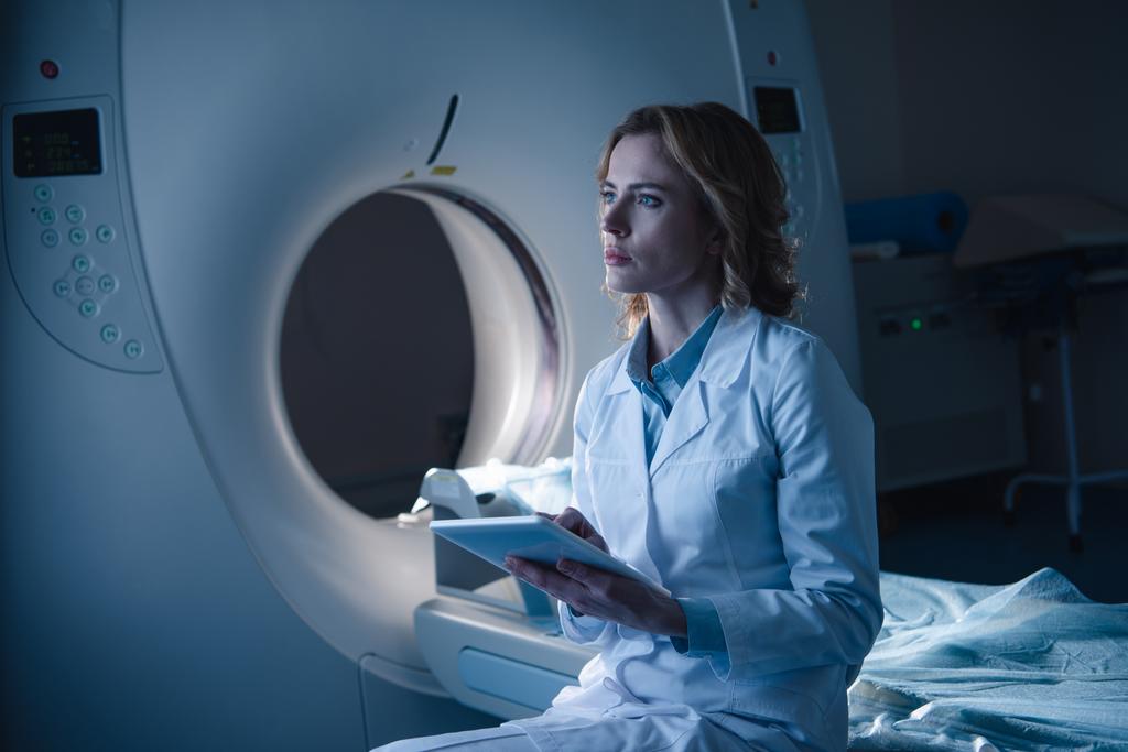 bilgisayarlı tomografi tarayıcı yakınında otururken ve uzağa bakarken dijital tablet tutan ciddi radyolog - Fotoğraf, Görsel