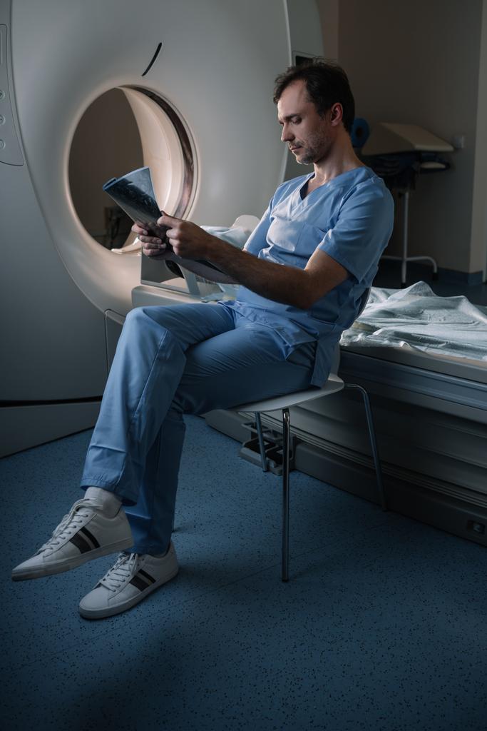 thoughtful doctor examining x-ray diagnosis while sitting near computed tomography scanner - Photo, Image
