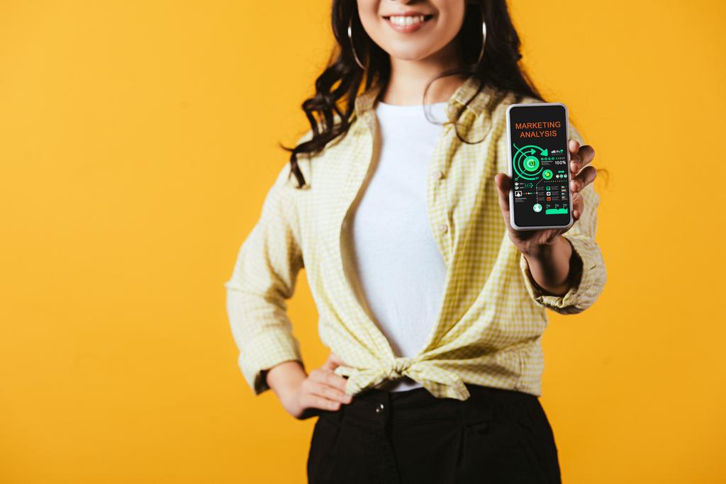 περικομμένη θέα της μελαχρινή κορίτσι που δείχνει το smartphone με ανάλυση μάρκετινγκ, απομονώνονται σε κίτρινο - Φωτογραφία, εικόνα