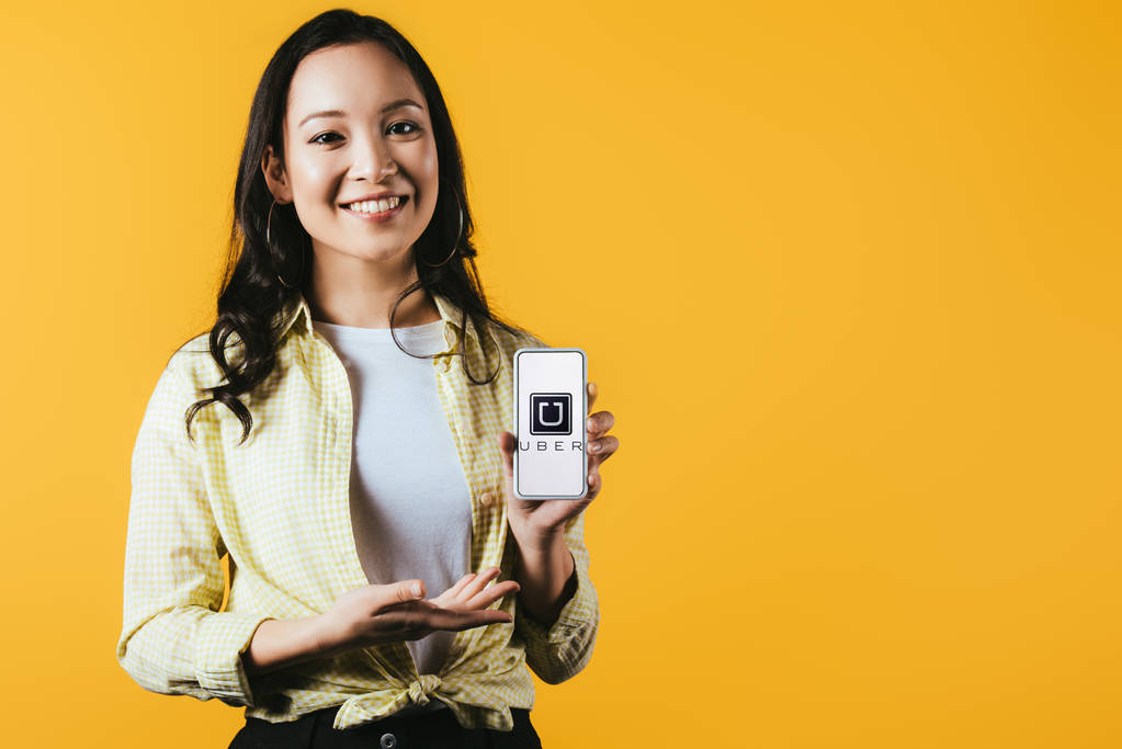 キエフ、 ウクライナ - 2019年4月16日:ユーバーアプリでスマートフォンを提示する笑顔のアジアの女の子、黄色で隔離 - 写真・画像
