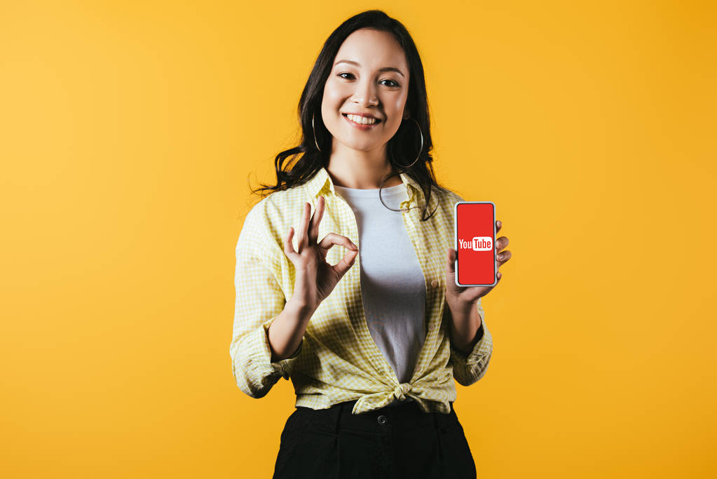 KYIV, UCRANIA - 16 DE ABRIL DE 2019: chica asiática sonriente mostrando signo de ok y teléfono inteligente con aplicación de youtube, aislado en amarillo
 - Foto, Imagen