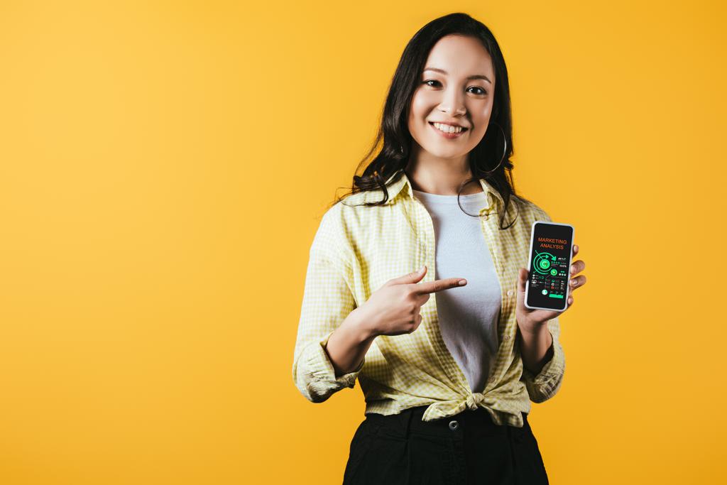 χαμόγελο ασιατικό κορίτσι δείχνει στο smartphone με ανάλυση μάρκετινγκ, απομονώνονται σε κίτρινο - Φωτογραφία, εικόνα