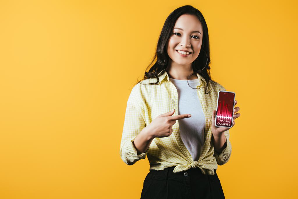 sorridente ragazza asiatica che punta a smartphone con corsi di trading, isolato su giallo
 - Foto, immagini