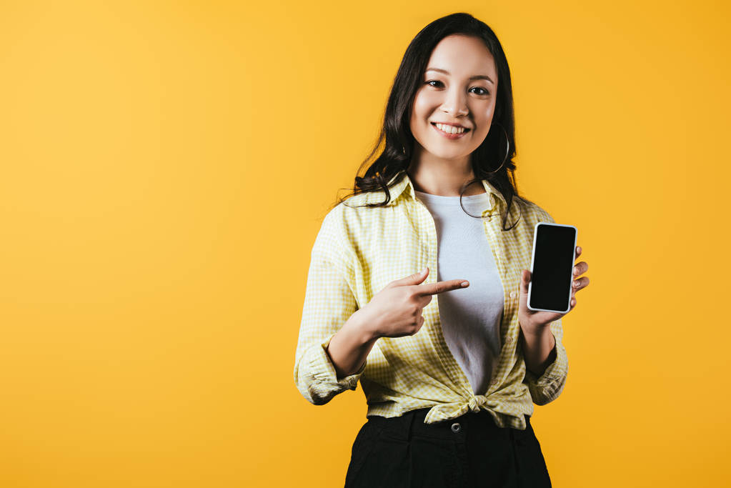 улыбающаяся азиатская девушка, указывающая на смартфон с пустым экраном, изолированным на желтом
 - Фото, изображение
