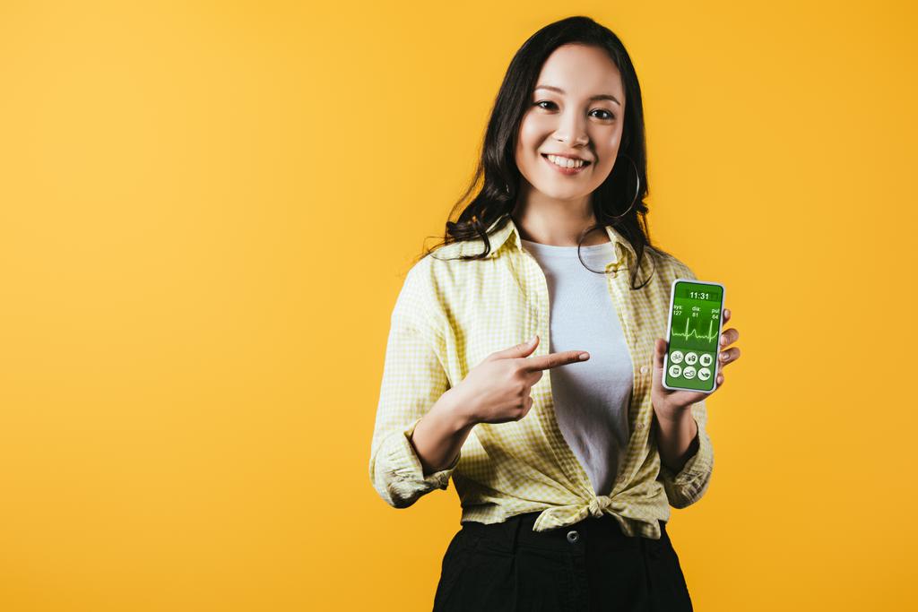 smiling asian girl pointing at smartphone with health app, isolated on yellow - Photo, Image