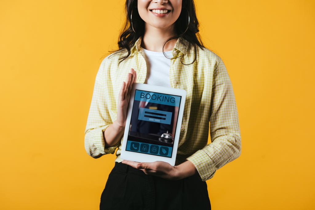 bijgesneden weergave van glimlachend meisje toont digitale tablet met Booking App, geïsoleerd op geel - Foto, afbeelding