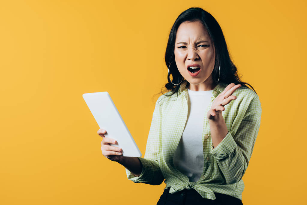 aggressive asian girl shouting and using digital tablet, isolated on yellow - Photo, Image