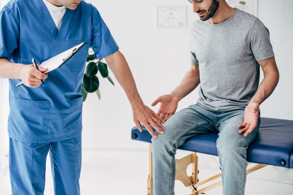 bijgesneden mening van arts die geduldige knie en holding Klembord in massage kabinet onderzoekt bij kliniek - Foto, afbeelding