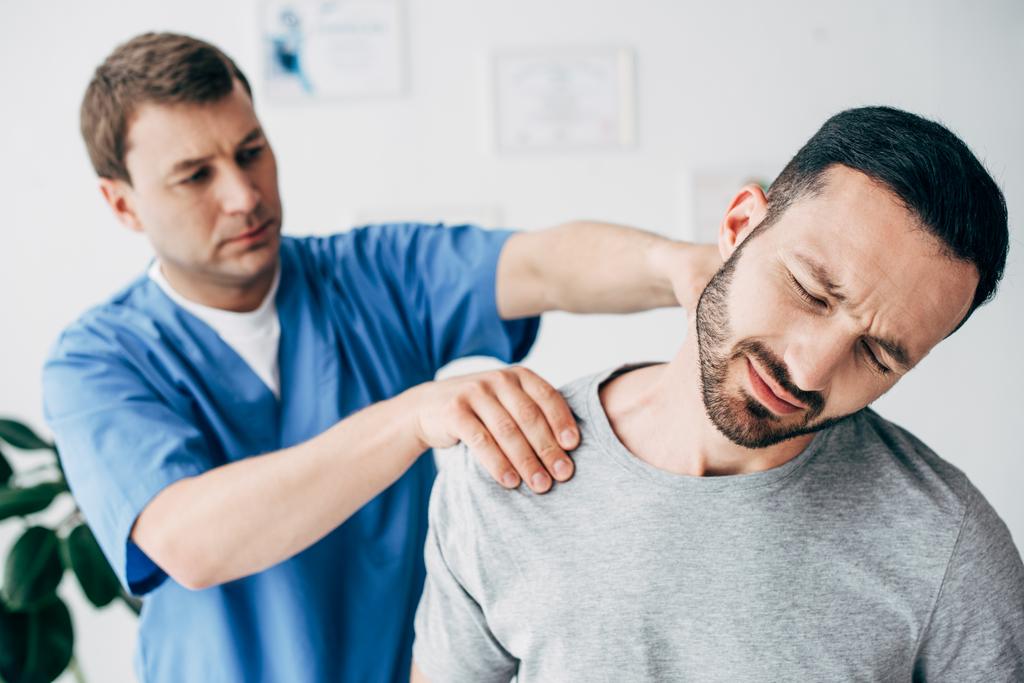 Enfoque selectivo del hombre que sufre y del doctor que masajea el cuello del hombre en gabinete de masaje en clínica
 - Foto, imagen