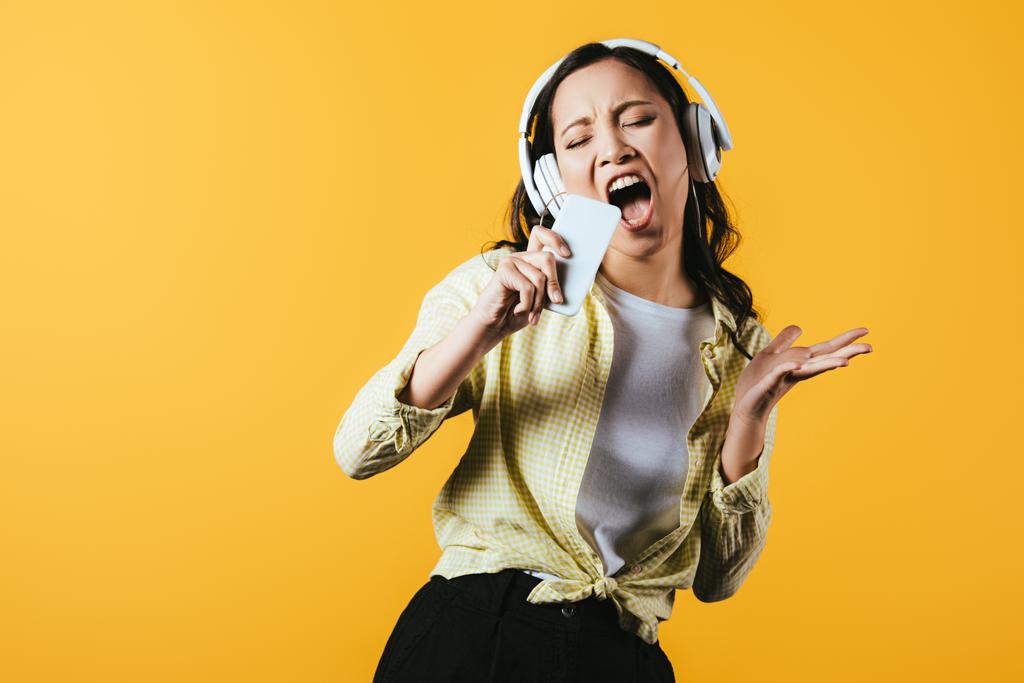 vonzó ázsiai lány éneklés és zenehallgatás zene fejhallgató és okostelefon, izolált sárga - Fotó, kép