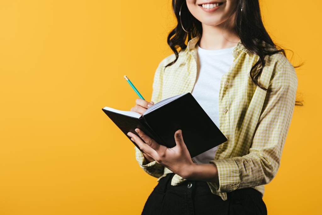 cropped view of smiling girl writing in notebook with pen, isolated on yellow  - Photo, Image