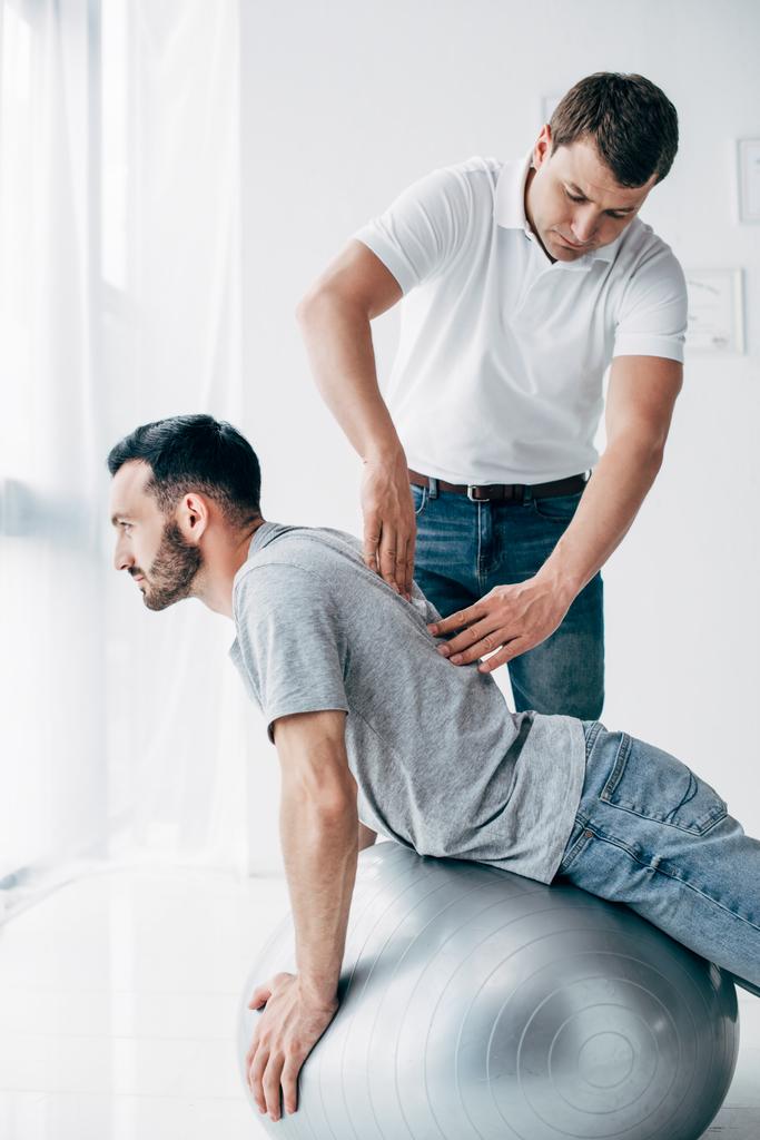 Chiropraktiker massiert Rücken eines gutaussehenden Mannes, der auf Fitnessball liegt - Foto, Bild