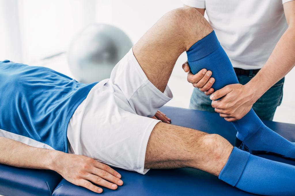 Περικομμένη θέα του φυσιοθεραπευτή μασάζ πόδι του παίκτη του ποδοσφαίρου στο νοσοκομείο - Φωτογραφία, εικόνα