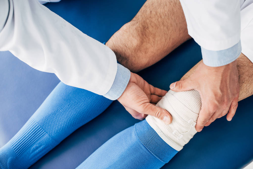Ausgeschnittene Ansicht des Physiotherapeuten, der elastischen Verband am Bein des Fußballers auf dem Massagetisch anlegt - Foto, Bild