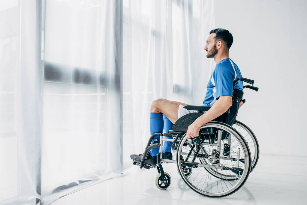 Mies jalkapallo univormu istuu pyörätuolissa ja katselee ikkunasta
 - Valokuva, kuva