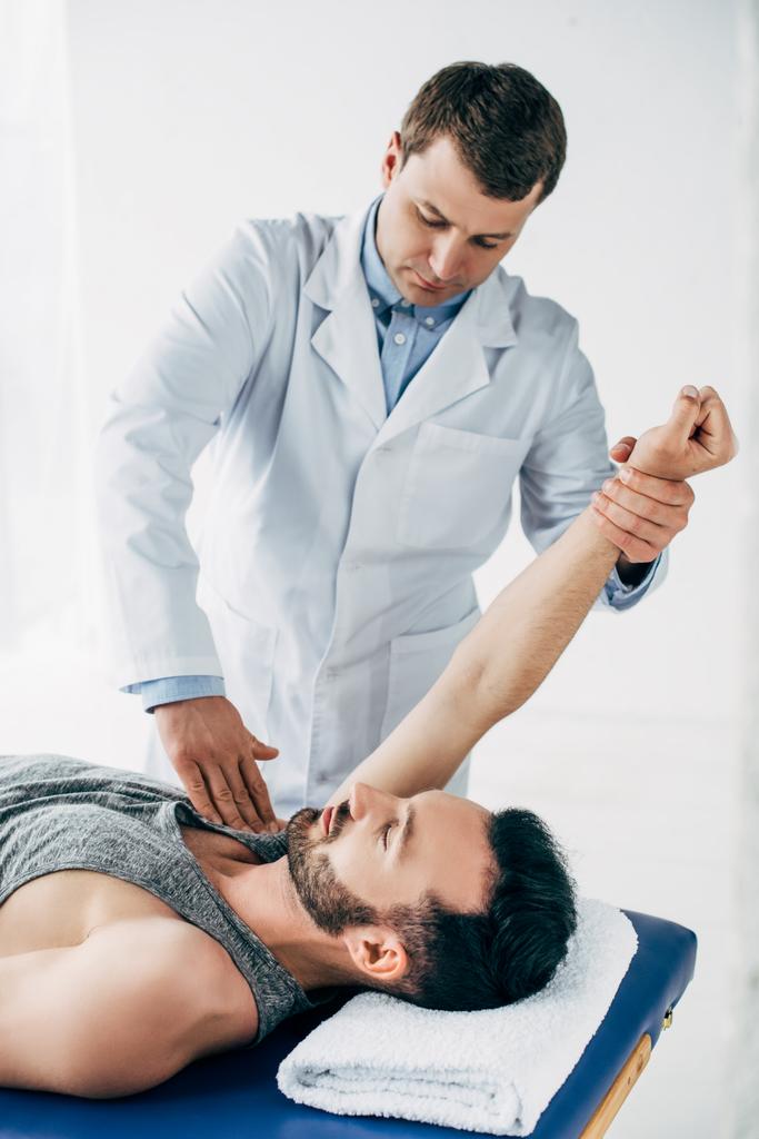 Chiropraktiker streckt Arm einer schönen Patientin aus, die mit Handtuch auf Massagetisch im Krankenhaus liegt - Foto, Bild