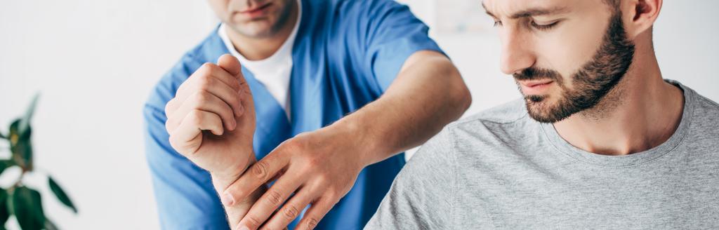 панорамный снимок мануального терапевта, массирующего руку бородатого пациента в больнице
 - Фото, изображение