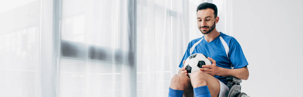 panoramische shot van de mens in het voetbal uniform zittend in een rolstoel en bedrijf Soccer Ball - Foto, afbeelding
