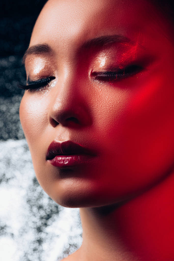 赤い光の中で目を閉じた魅惑的なアジアの女の子のクローズアップ  - 写真・画像