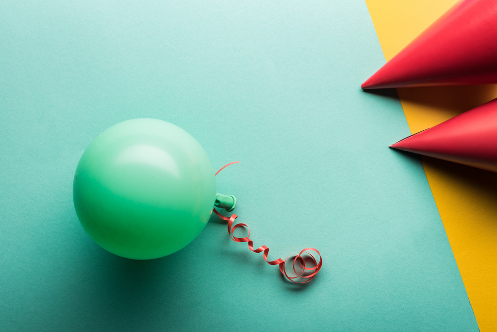 Draufsicht auf grünen Ballon und rote Party-Hüte auf tiffanischem und gelbem Hintergrund - Foto, Bild