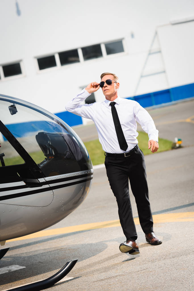 bonito piloto no formal desgaste ajustando óculos de sol e andando perto de helicóptero
 - Foto, Imagem