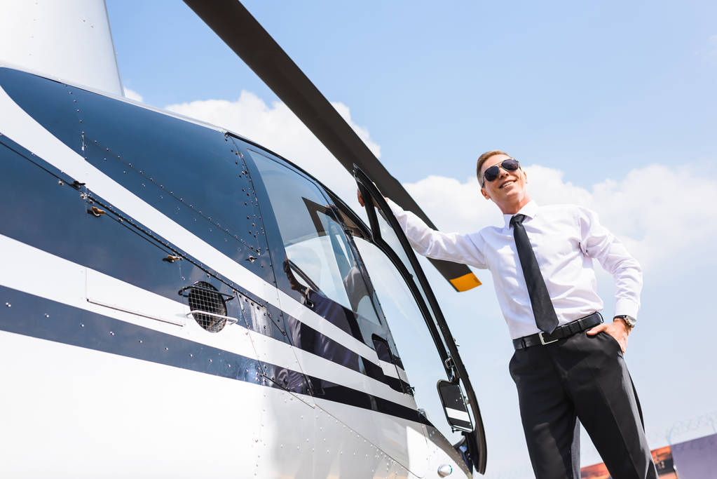 schöner Pilot in offizieller Kleidung und Sonnenbrille, der in der Nähe des Hubschraubers lächelt - Foto, Bild