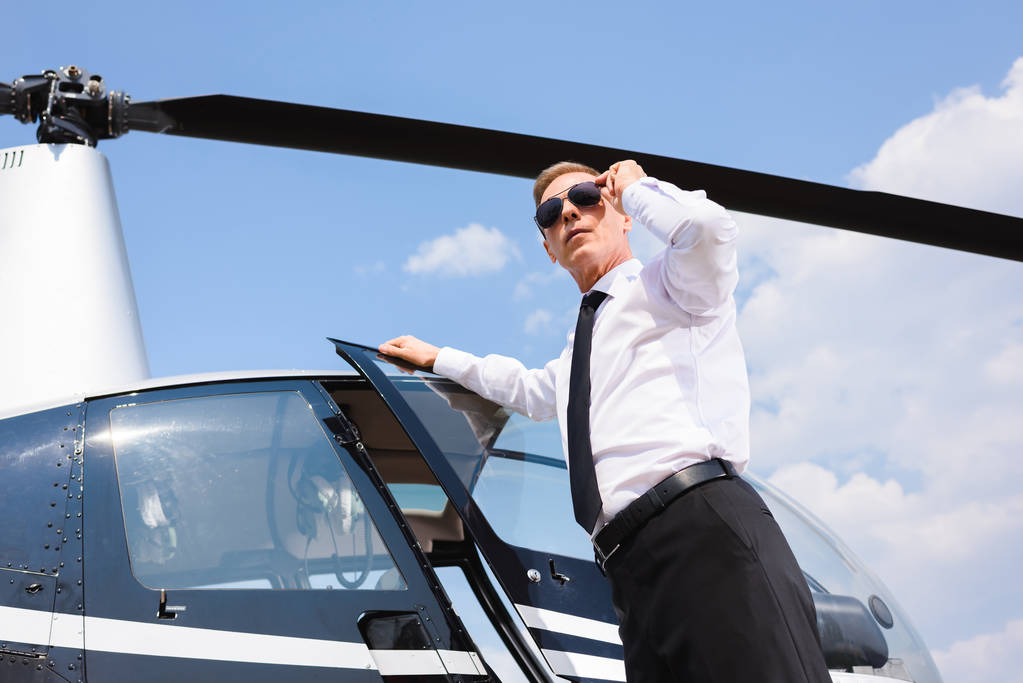 pohled nízkého úhlu pilota ve formálním opotřebení a slunečních brýlích poblíž vrtulníku - Fotografie, Obrázek